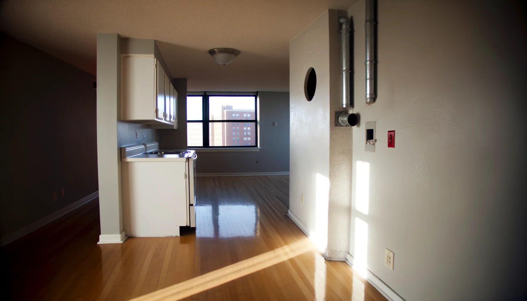 A spacious, empty Kansas City apartment kitchen and laundry area with exposed hookups and connections visible, showing the stark white or light-colored walls, polished hardwood or tile flooring, and d